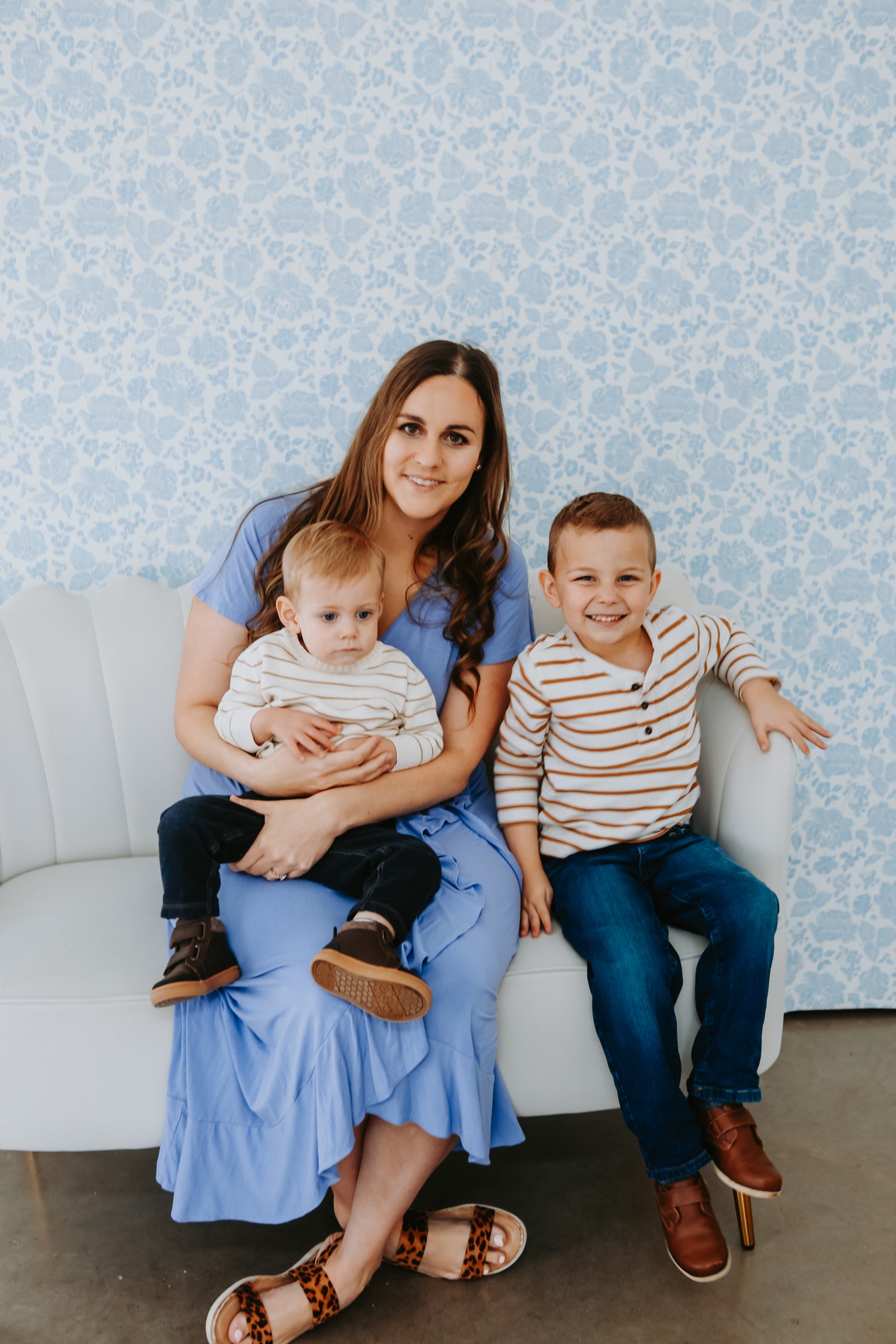 Woman in a blue dress sitting on a white couch with two children against a blue patterned wall.