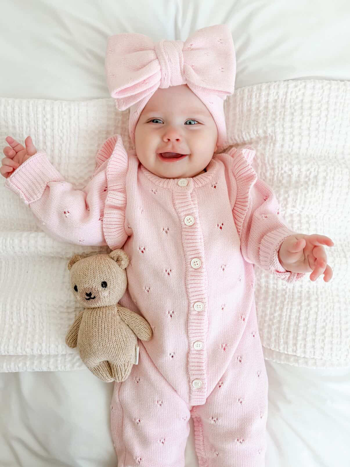 Baby in pink outfit with a bow and teddy bear on a white blanket