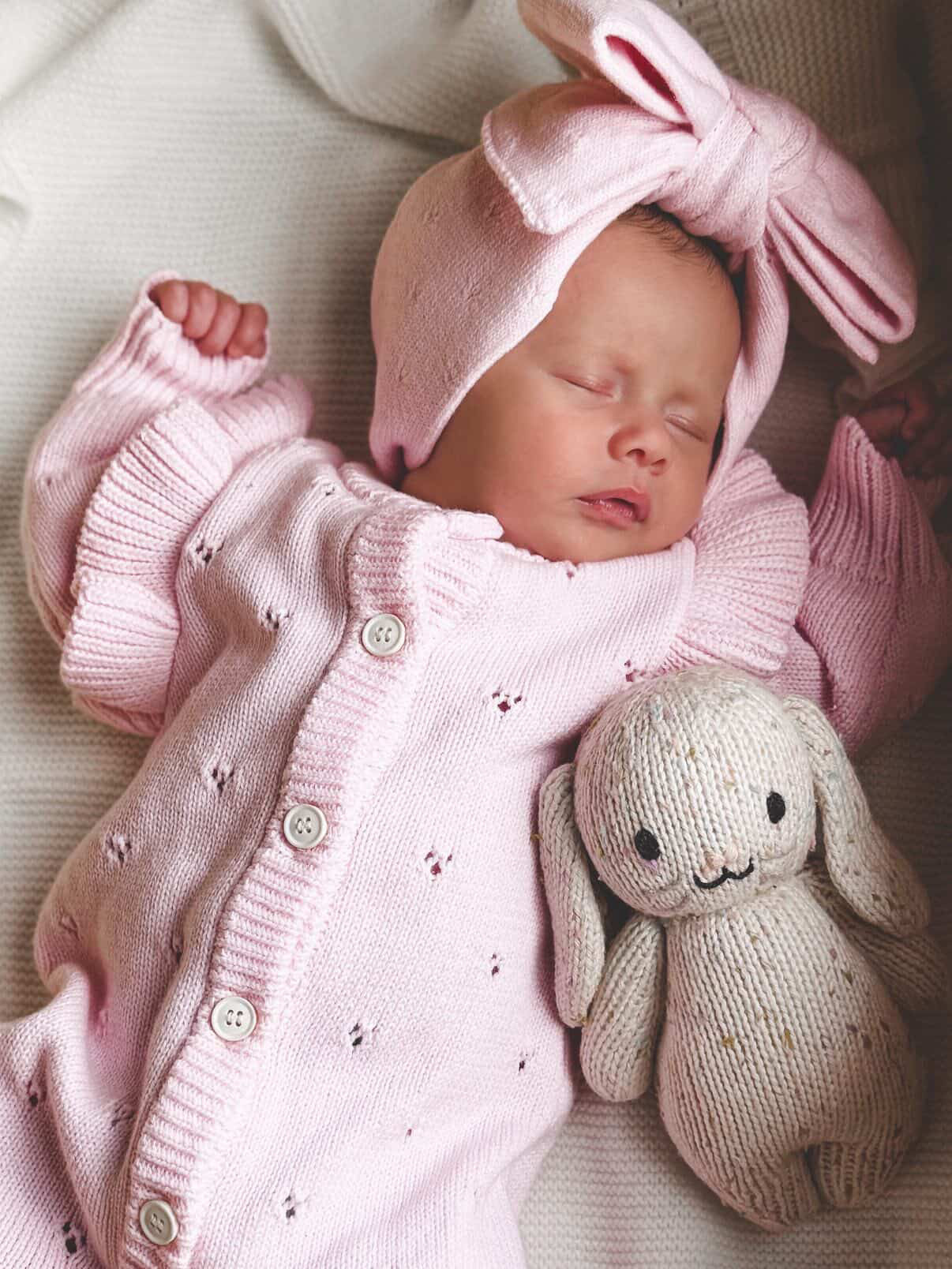 Newborn baby in pink outfit with bunny ears and toy, sleeping peacefully.