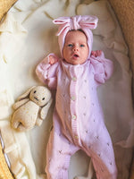 Baby in a pink outfit with a bow headband lying on a soft surface next to a plush toy.