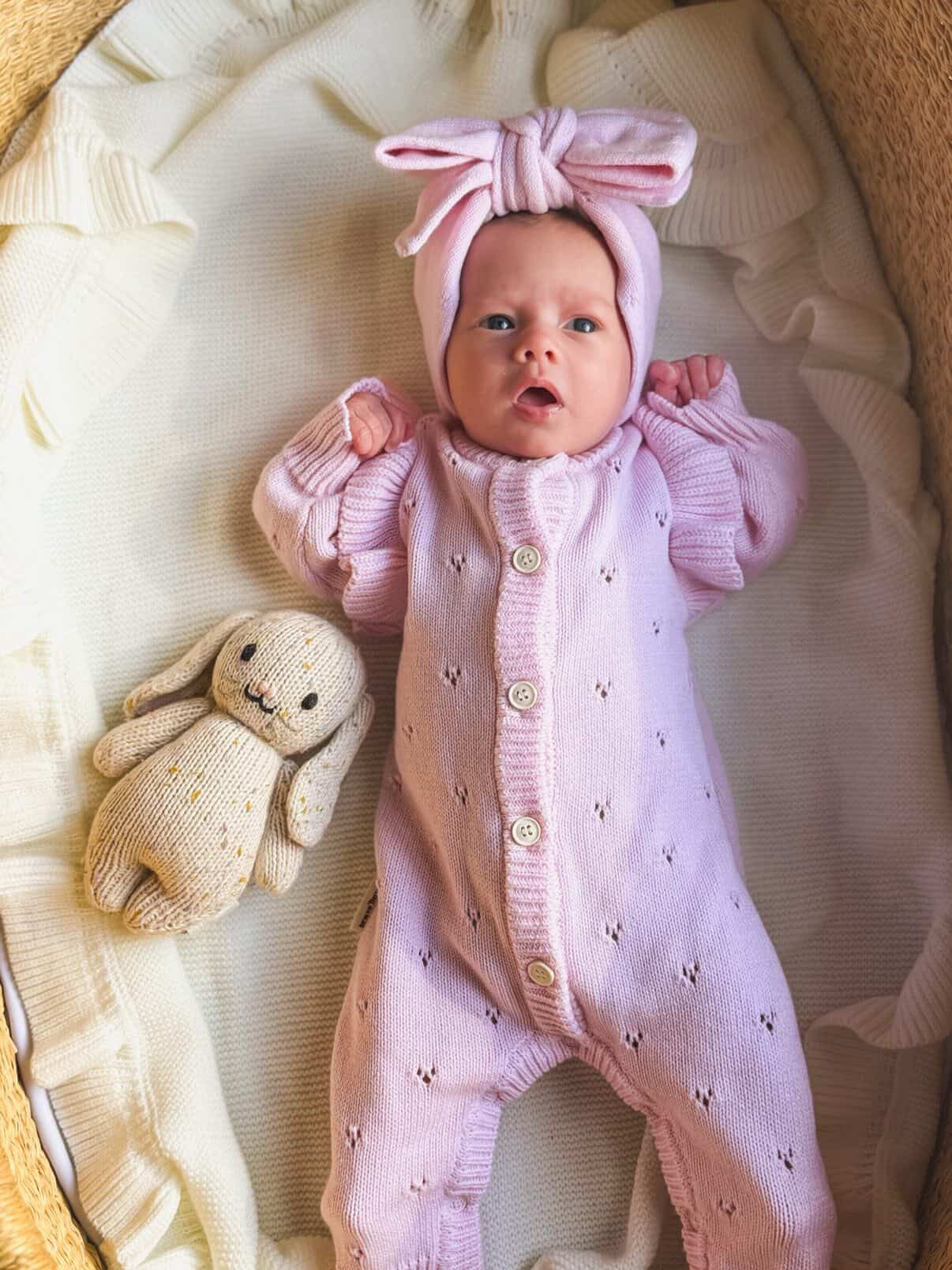 Baby in a pink outfit with a bow headband lying on a soft surface next to a plush toy.