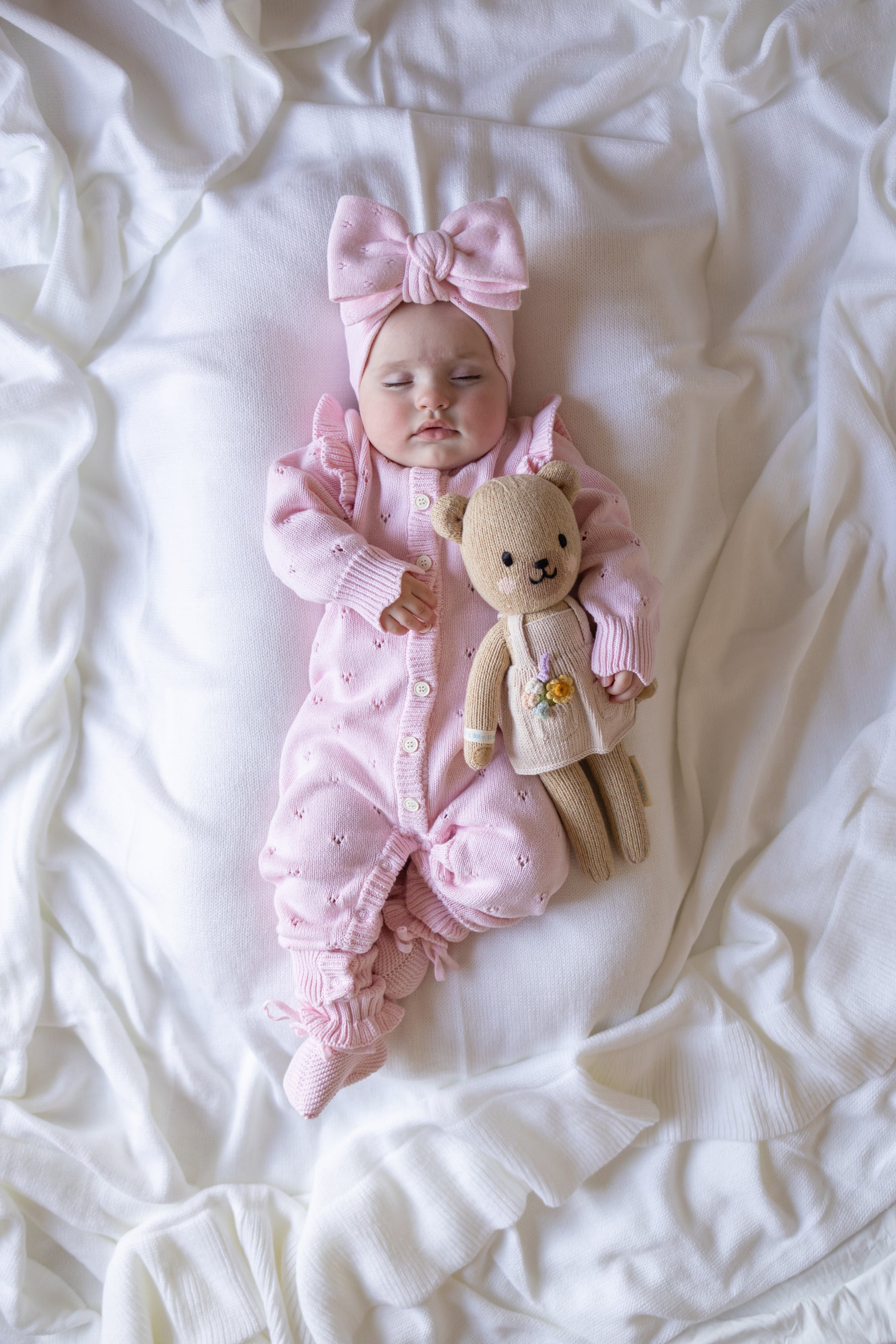 Baby in pink outfit with a teddy bear on a white blanket