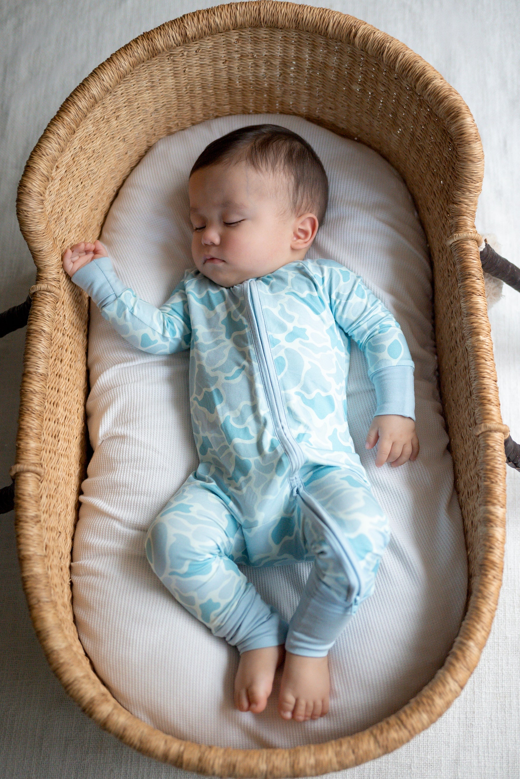 Baby in a blue onesie with cloud pattern lying in a wicker crib.