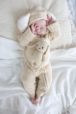 Baby in a beige knitted outfit with a hood and bear plushie, lying on a white blanket.