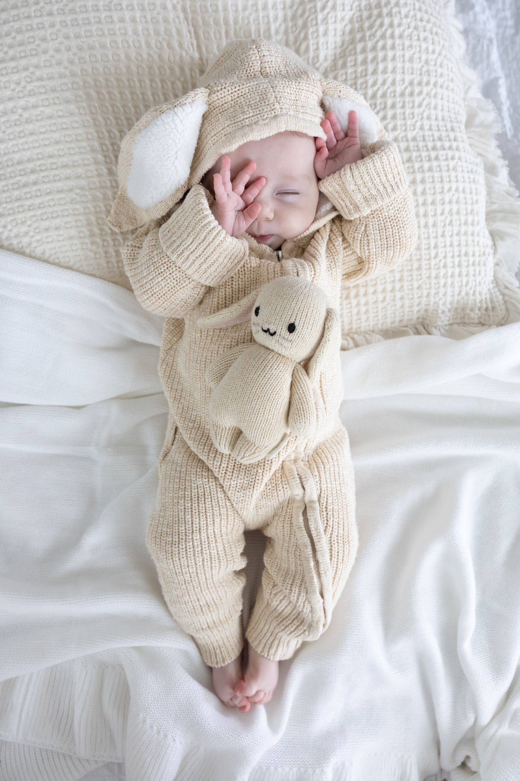 Baby in a beige knitted outfit with a hood and bear plushie, lying on a white blanket.
