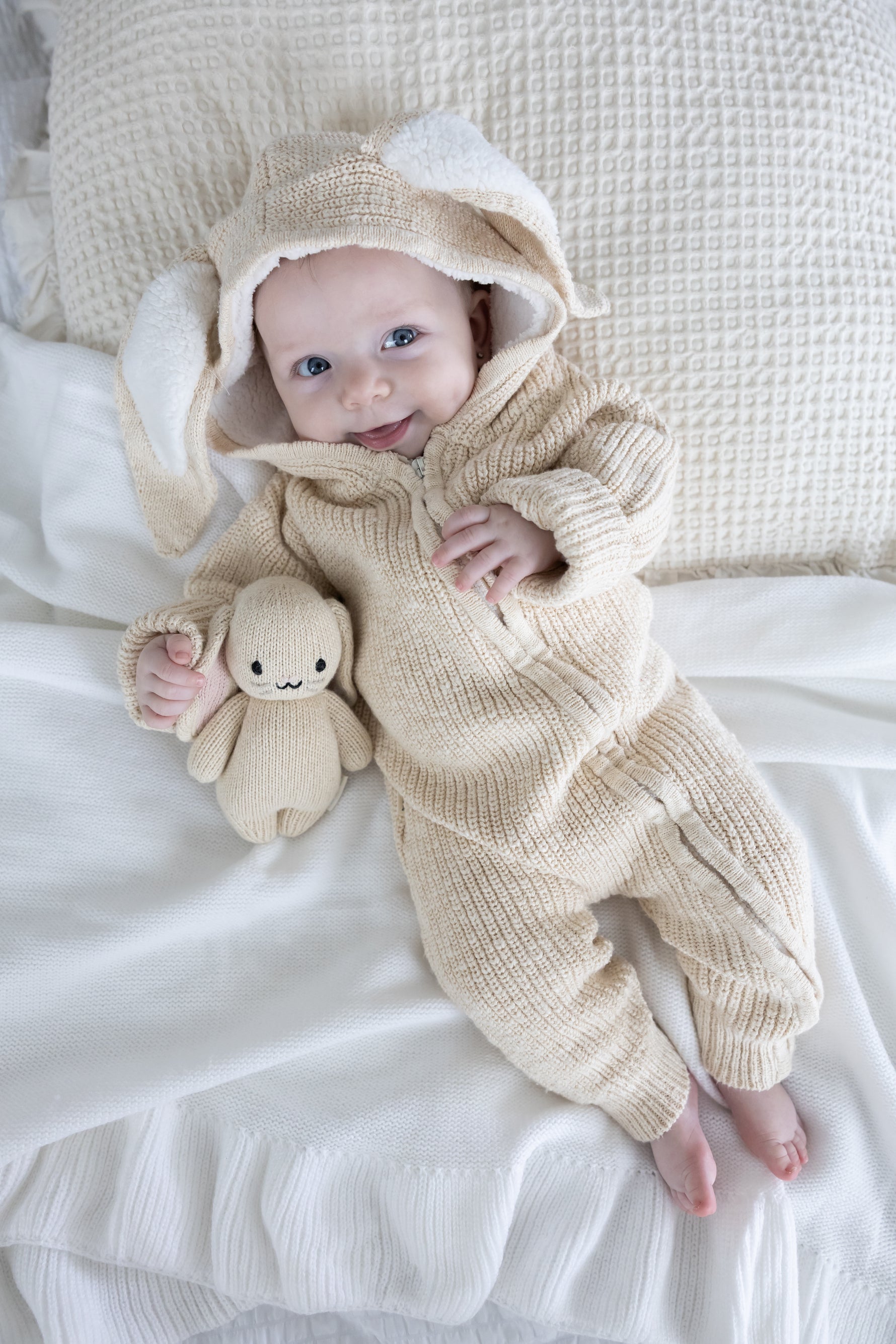 Baby in a beige knitted outfit with a hood and bunny ears, holding a plush toy, on a white blanket.
