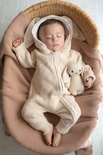 Baby in a beige knitted outfit holding a plush toy, lying on a brown blanket.
