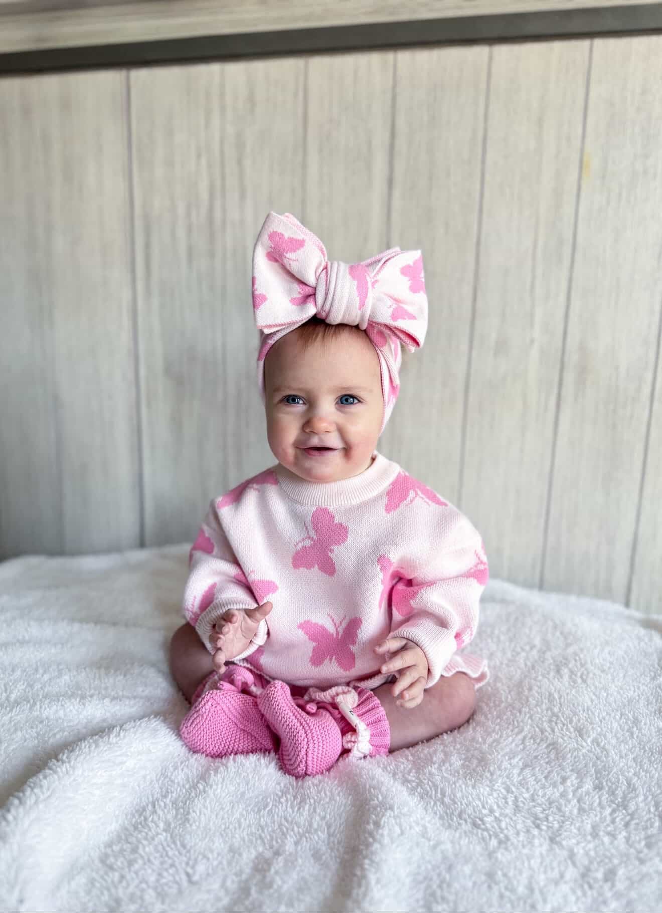 Baby wearing a pink outfit with butterfly designs and a matching headband, sitting on a white blanket.