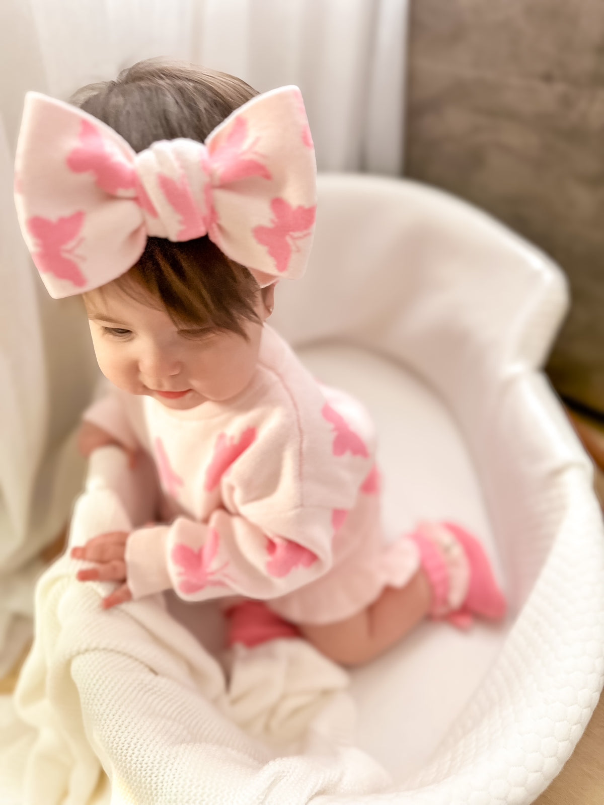Baby in a pink outfit with a large bow sitting on a white blanket.