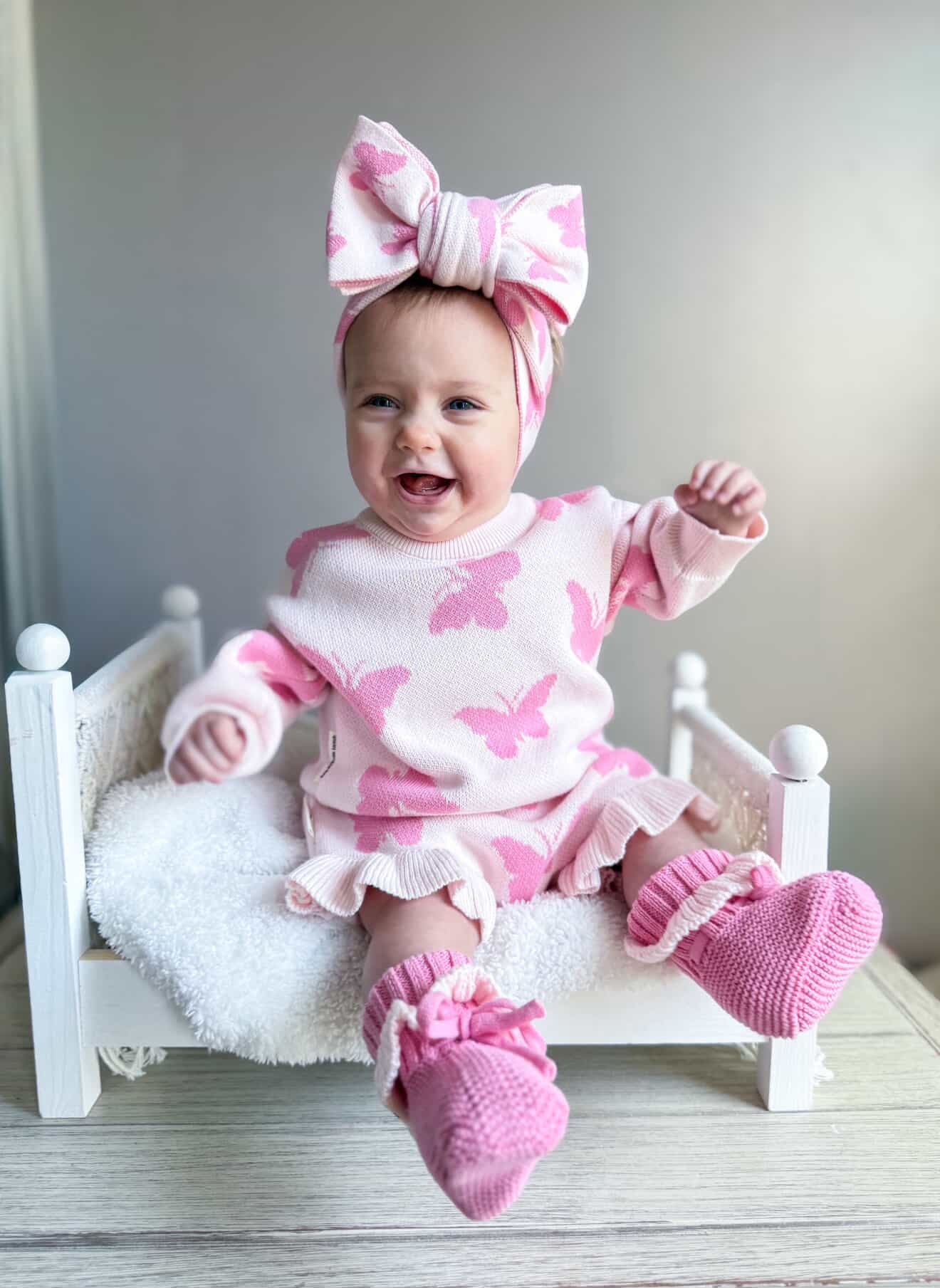 Baby in pink butterfly outfit with matching headband sitting on a small white bed.