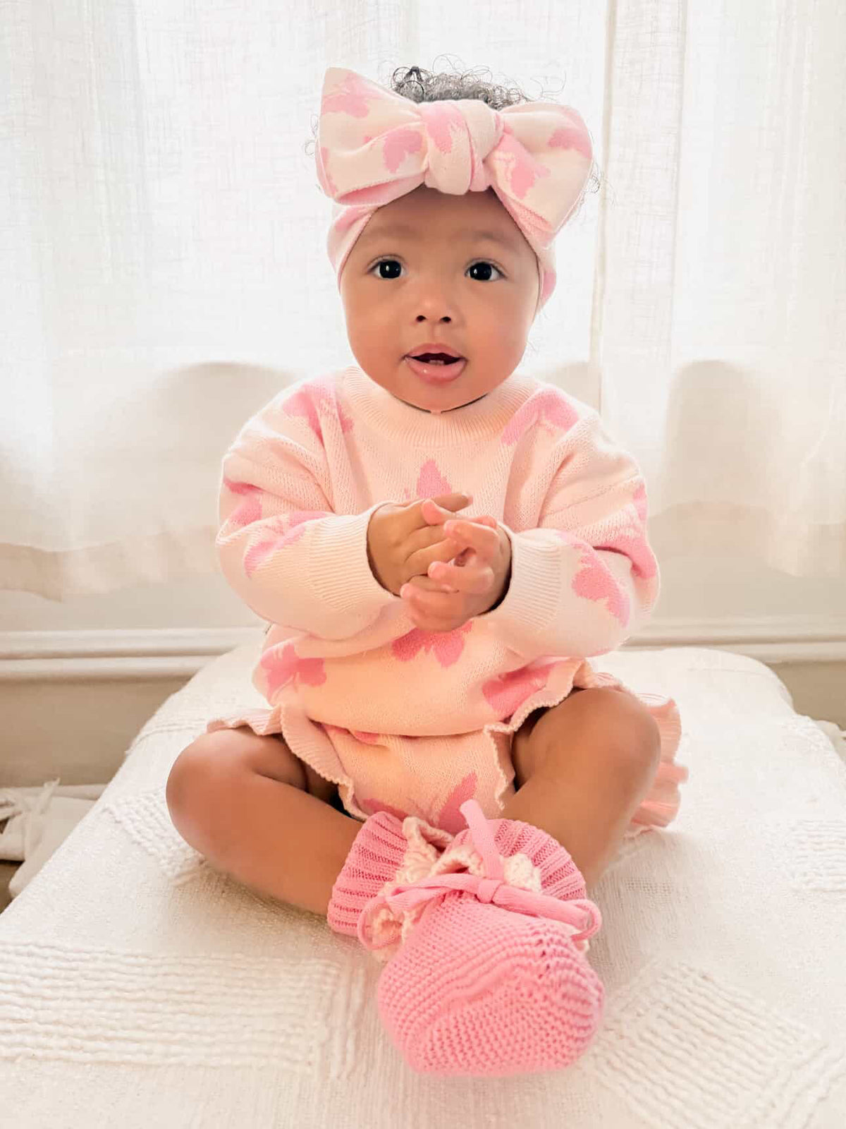 Baby in pink outfit with matching headband and booties sitting on a white surface.