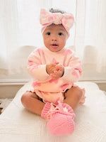 Baby in pink outfit with matching headband and booties sitting on a white surface.