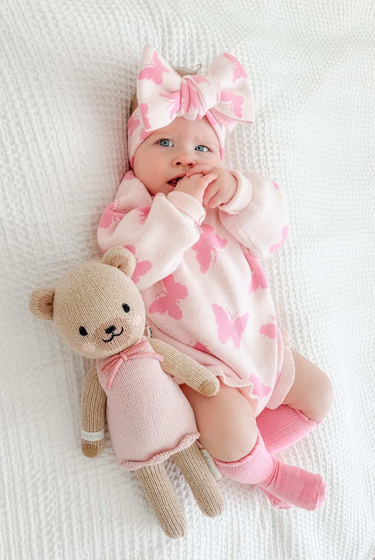 Baby in pink floral outfit with teddy bear toy on white blanket