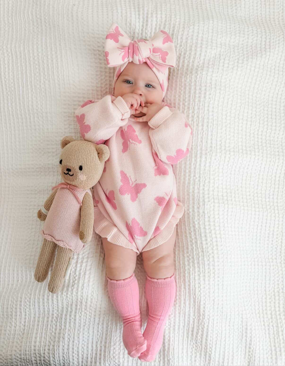Baby in pink outfit with butterfly patterns, holding a teddy bear, on a white blanket.