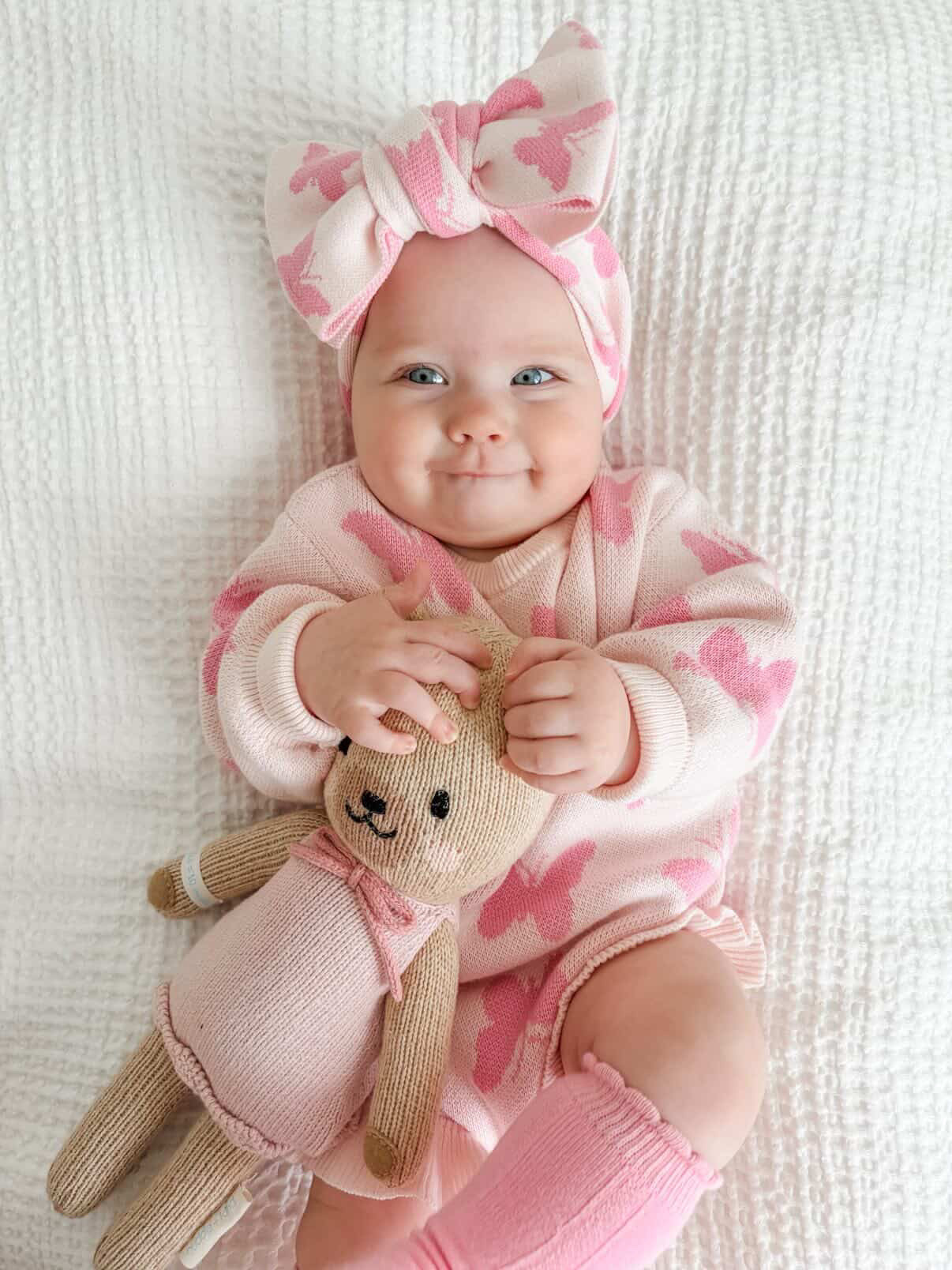 Baby in pink outfit with a bow, holding a teddy bear on a white blanket