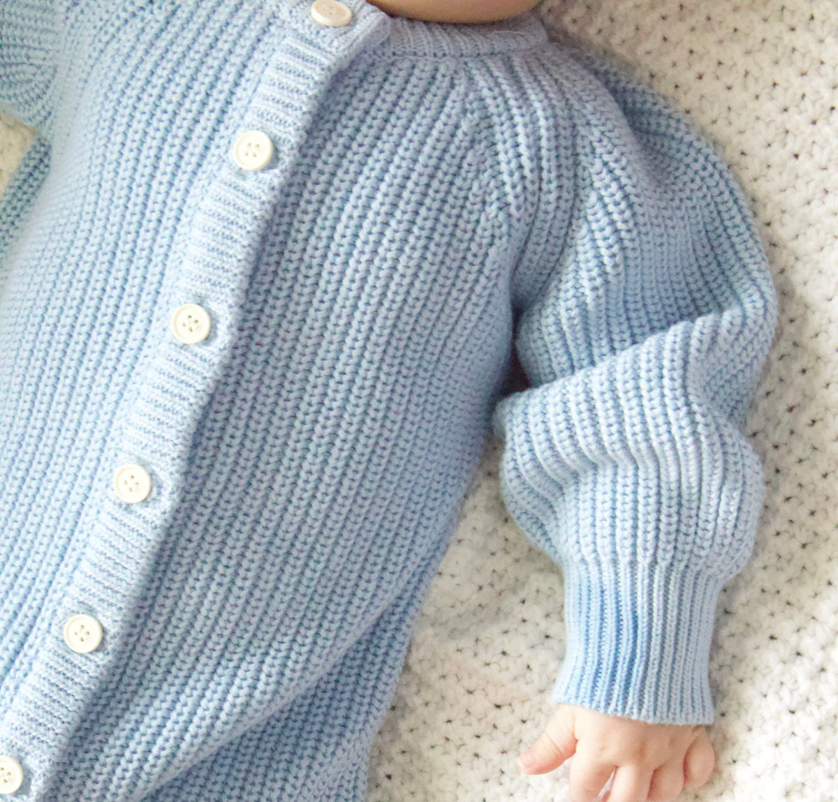 Close-up of a baby in a light blue knitted outfit with buttons on a white blanket.