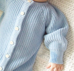 Close-up of a baby in a light blue knitted outfit with buttons on a white blanket.