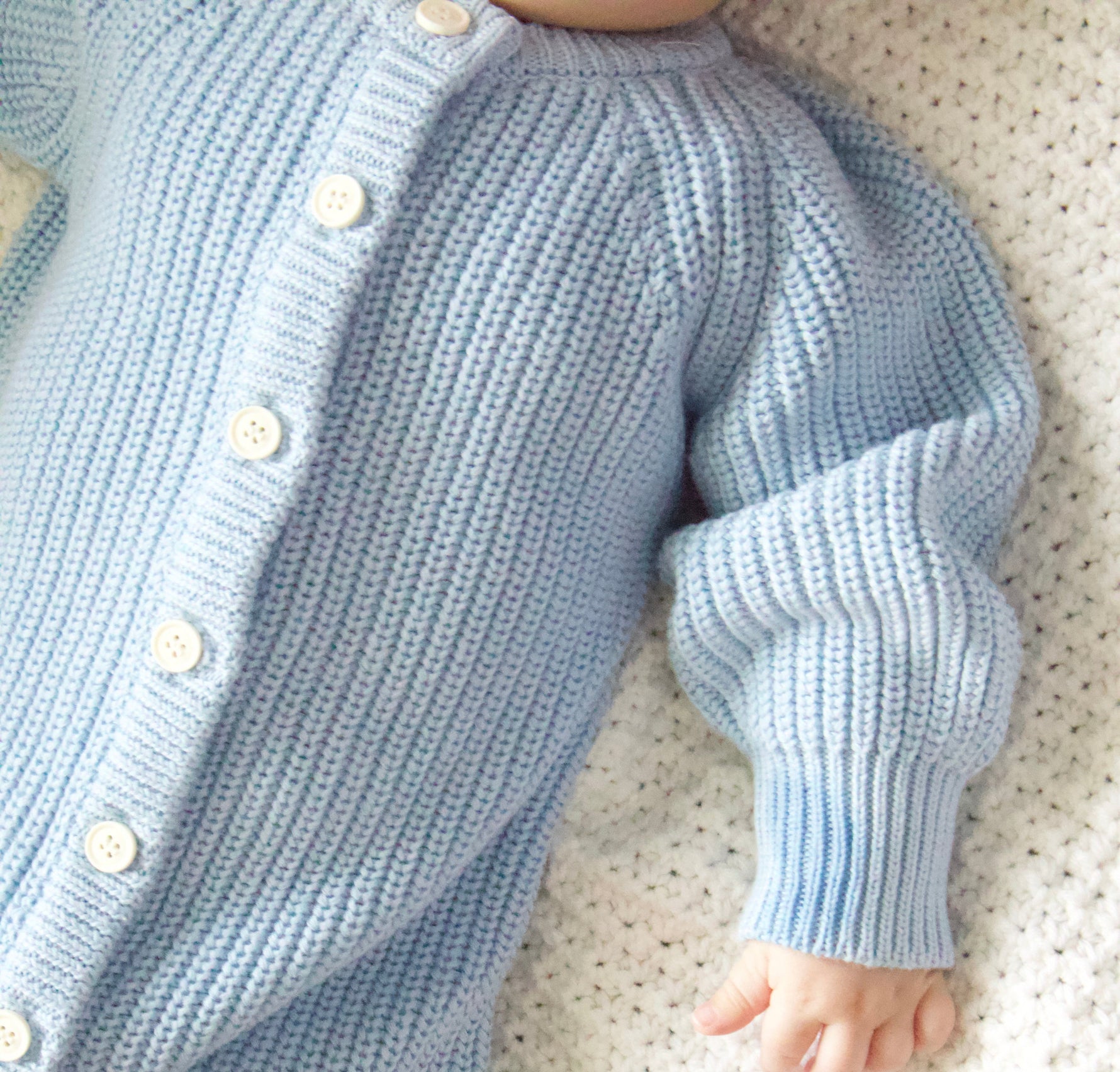 Close-up of a baby in a light blue knitted outfit with buttons on a white blanket.
