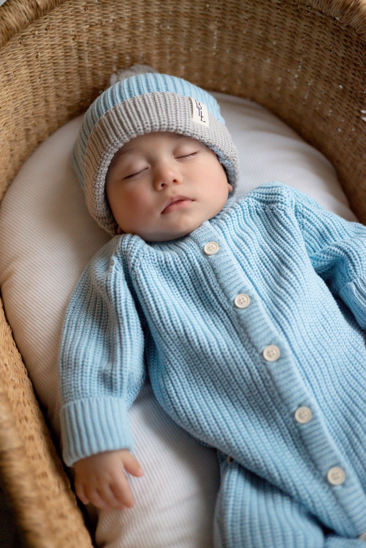 Baby in a blue knitted outfit and hat sleeping in a woven basket.
