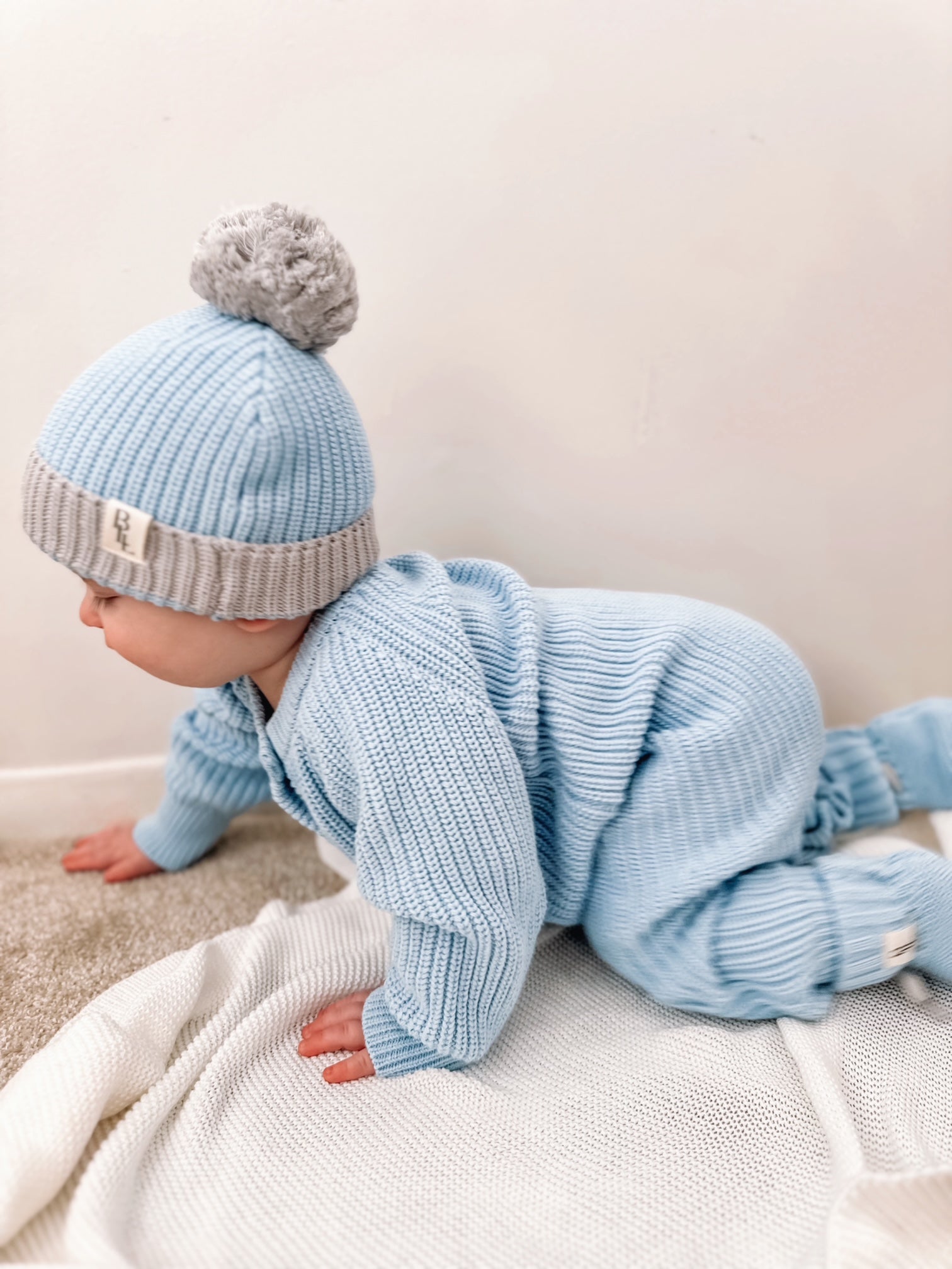 Baby in light blue knitted outfit and hat with pom-pom on a white blanket