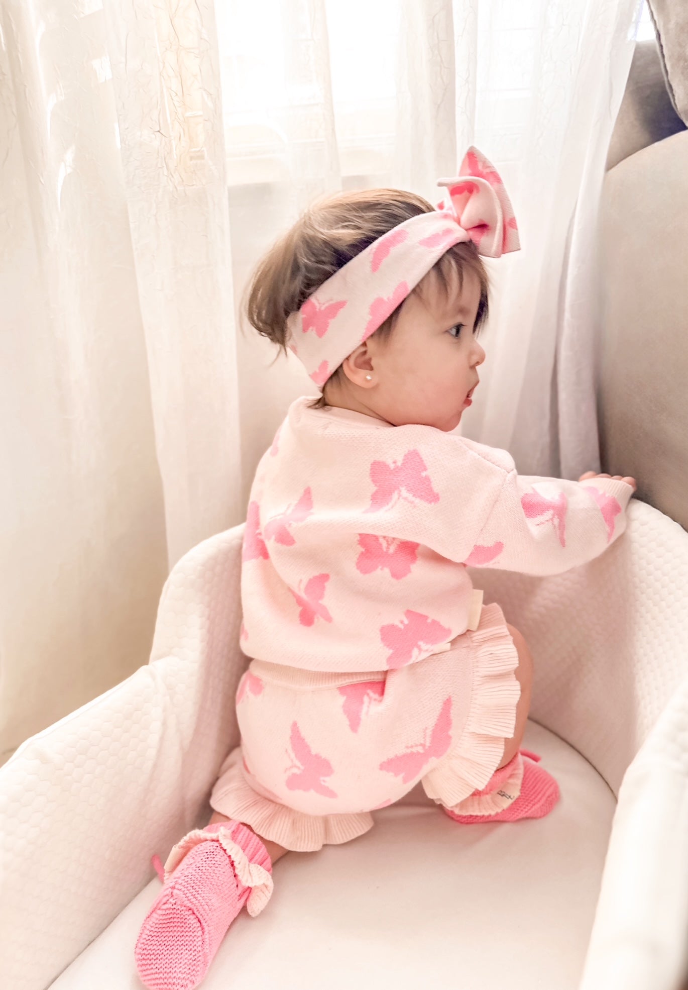 Baby in pink butterfly outfit sitting on a white chair with white curtains in the background