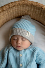 Baby wearing a blue and gray knit hat and sweater, sleeping in a wicker basket.