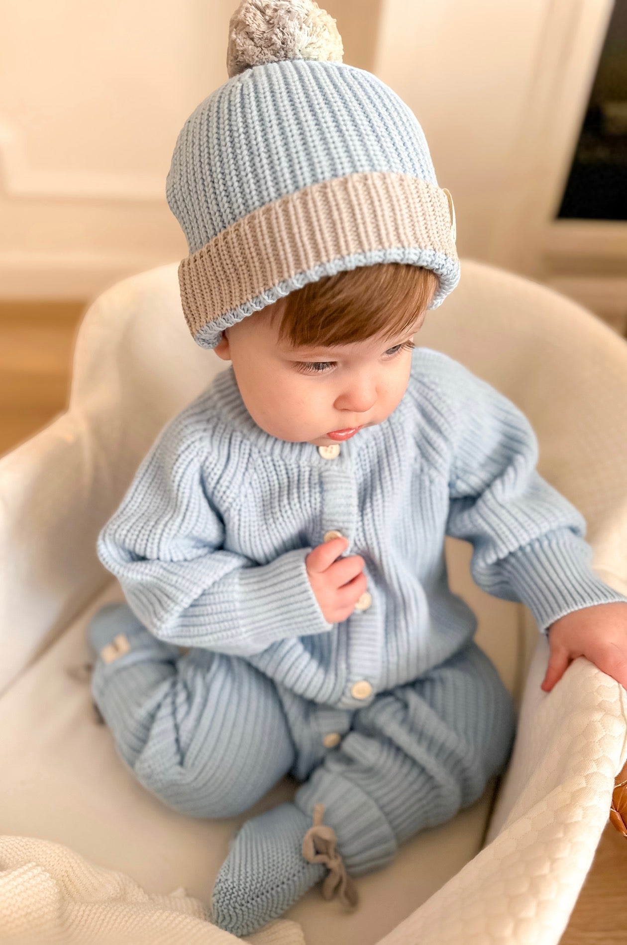 Baby wearing a light blue knitted outfit with a matching hat, sitting in a white chair.