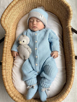 Baby in a blue knitted outfit with a matching hat, lying in a crib with a teddy bear.
